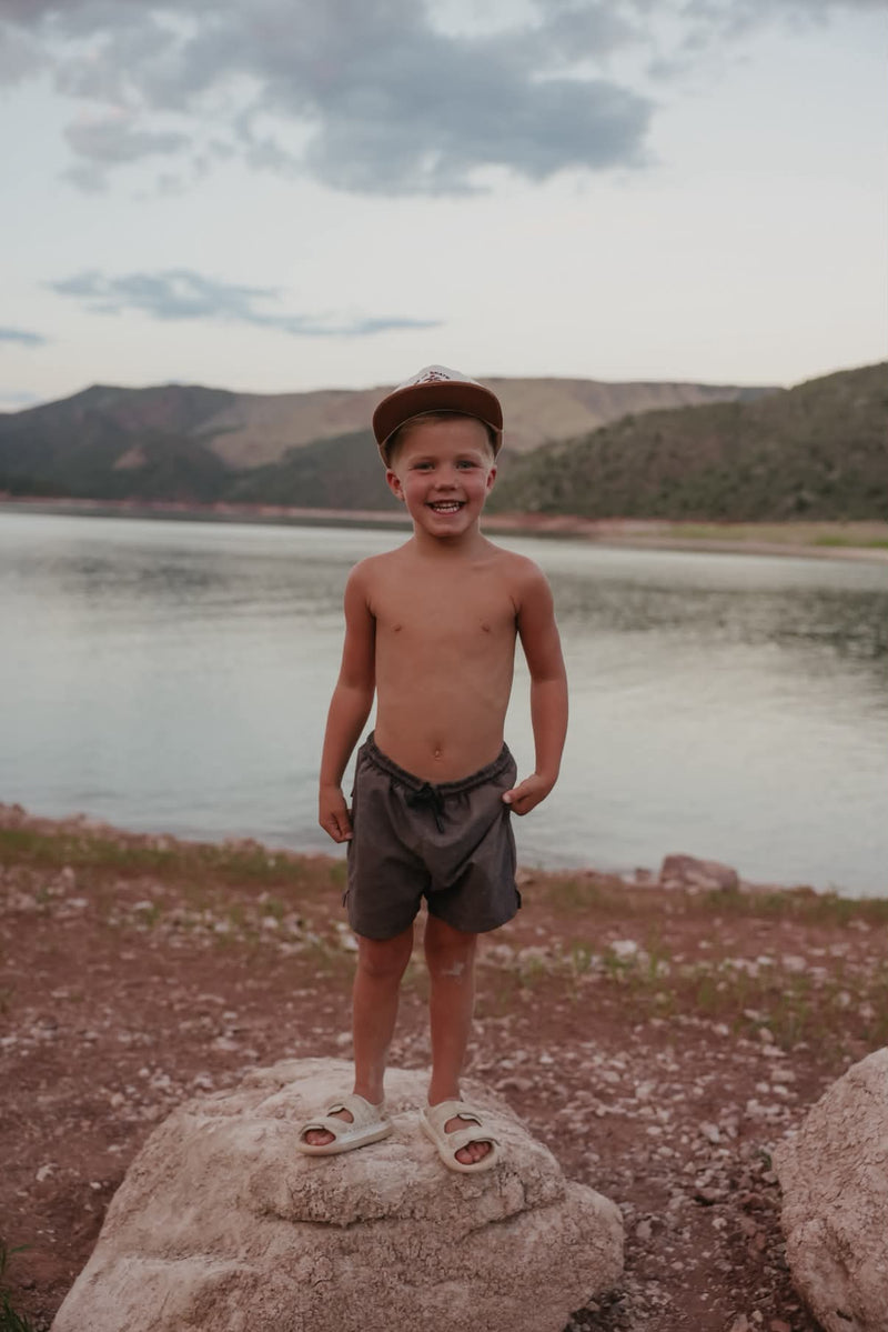 Boy wearing charcoal palm shadow swim shorts standing on a rock beside a mountain lake