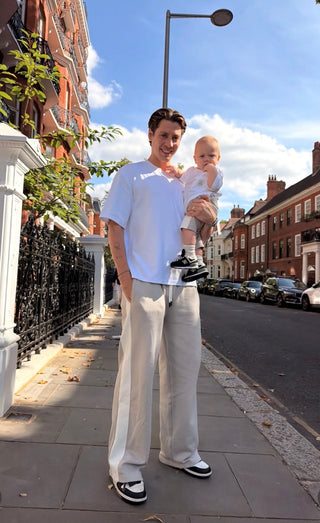 Sam Vanderpump holding his baby while wearing matching white MANCUB T-shirts on a city street