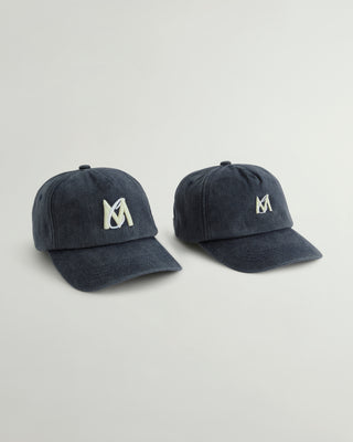 Two navy blue baseball caps with a white embroidered stylized 'M' logo on the front, displayed side-by-side on a light gray background.